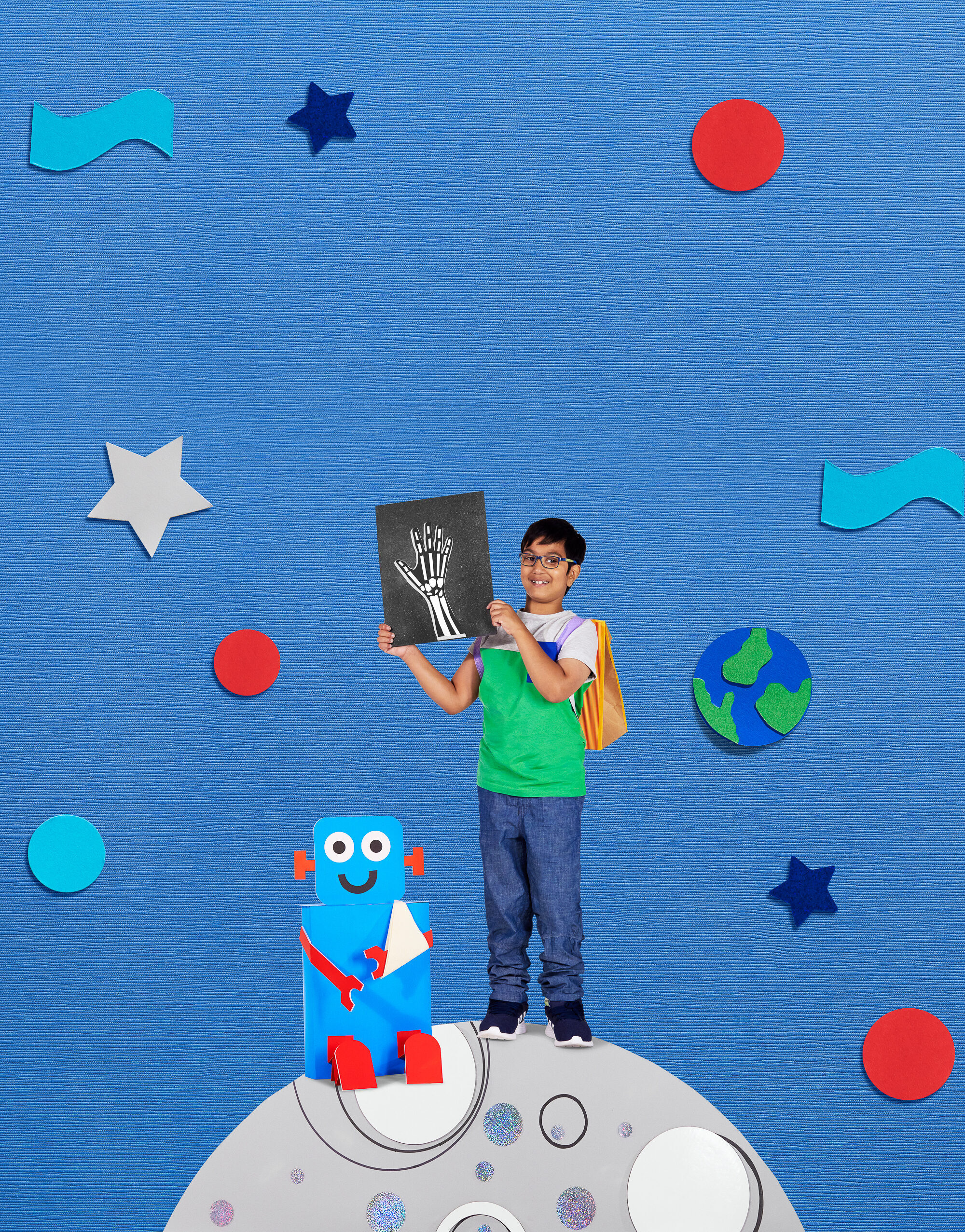 A boy with medium skin tone and dark hair, wearing a backpack and holding up an x-ray of a hand. He is standing on a cut-out image of a moon with a blue robot sitting next to him.