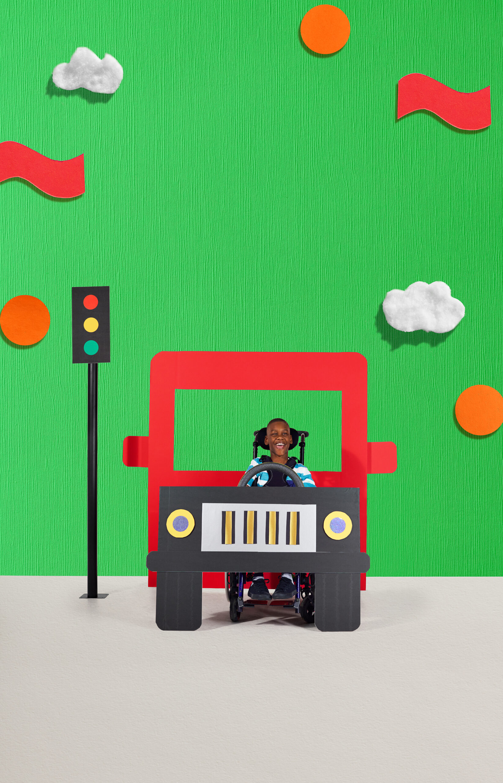 A boy with dark skin tone in a wheelchair, behind the wheel of a red bus.