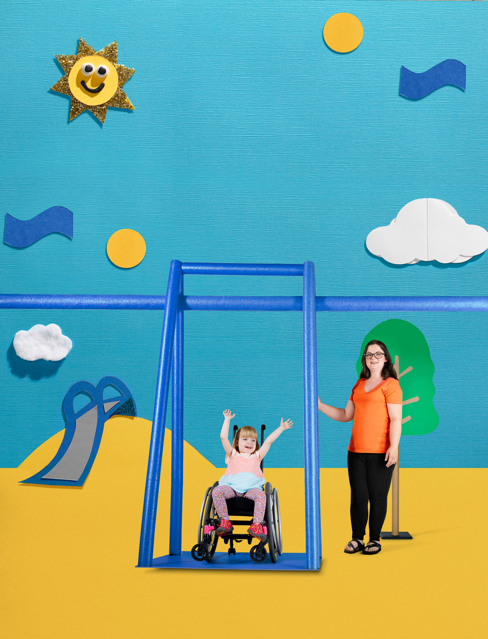 A child with light skin tone and blonde hair using a wheelchair on an accessible swing at the playground. She is waving her arms in the air. She is next to her mother, who has light skin tone and brown hair. She is wearing an orange shirt and black pants.