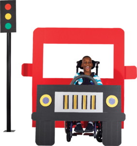 A boy with dark skin tone in a wheelchair, behind the wheel of a red bus.