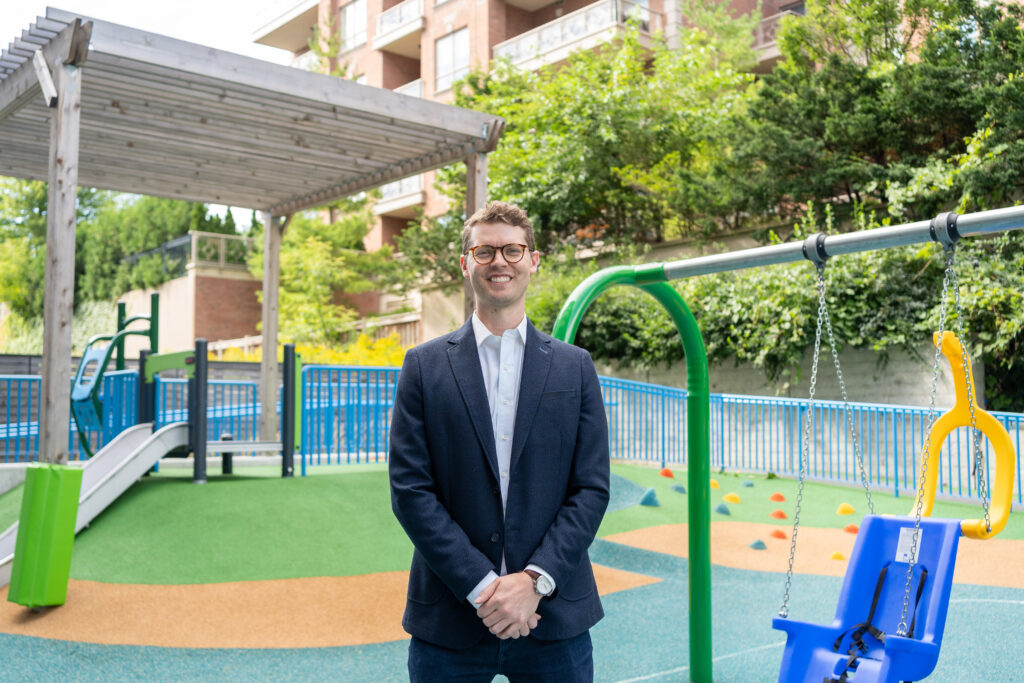 An adult with light skin tone and short brown hair. He is wearing glasses, a white dress shirt and a blue suit jacket. He is standing in front of the accessible slides and swings on the playground at Holland Bloorview.